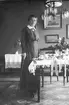 Sigrid Paues stående vid ett fikabord. Interiörsbild. Tidsomfånget är 1900 - 1940