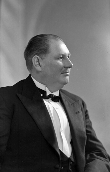 Foto av en medelålders man i smoking (?) frack (?) med mörk fluga och väst.Midjebild, halvprofil. Ateljéfoto.