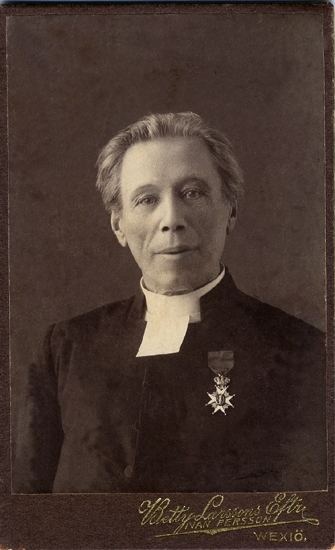 Porträtt (bröstbild, halvprofil) av en äldre man i prästrock och
prästkrage. På bröstet syns Vasa-orden. 

Claes Aug. Lind -  (1849-1933). Student i Växjö 1872. 
Senare kyrkoherde och prost i Älmeboda, Växjö stift, Kronobergs län, fr. 1898-   . 
Källa: Svensk Rikskalender 1908, s. 438."Lärare och studenter vid Växjö Högre Allmänna Läroverk 1850-1950", (Växjö 1951) s. 66.