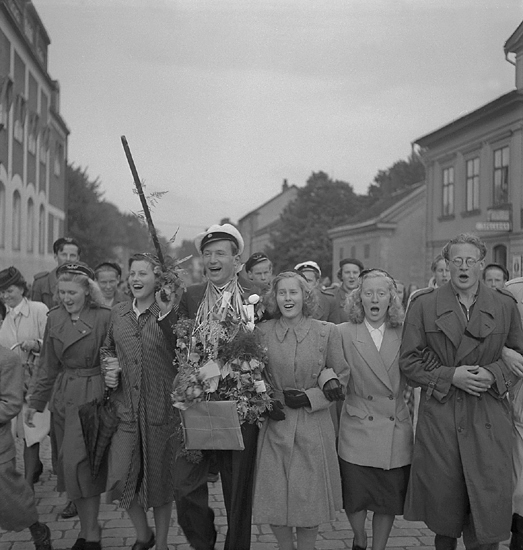 Studenterna, 28/5 1945. 
En glad student med anhöriga och vänner på väg upp till Stortorget, efter 
att ha lyssnat till talet vid Esaias Tegnérs staty. I bakgrunden skymtar bl a Smålandspostens hus.

Vad gäller avgångsklasserna 1945 - Se "Lärare och Studenter vid
Växjö Högre Allmänna Läroverk 1850-1950" (1951), s. 177-180.