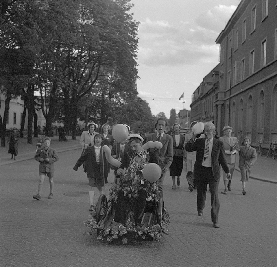 Studenterna andra dagen, 1950.
En student körs i rullstol uppför Kungsgatan. Till höger syns dåv. Sparbankshuset på kv. Sunaman 2. 
Något suddigt foto. 
Angående avgångsklasserna 1950 - se "Lärare och Studenter vid Växjö 
Högre Allmänna Läroverk 1850-1950" (1951), s. 193-196, 289.