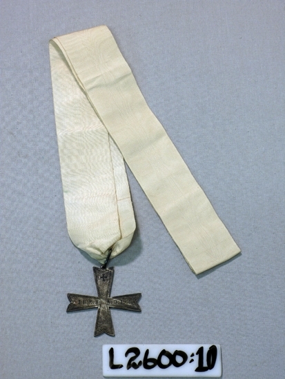 Ett 40 mm brett vitt ripsband med vidhängande malteserkors i 
vitmetall.
Text: IBMBADIC

Inskrivet i huvudboken 1933.
Funktion: Ordenstecken (inom Frimurarorden ?)