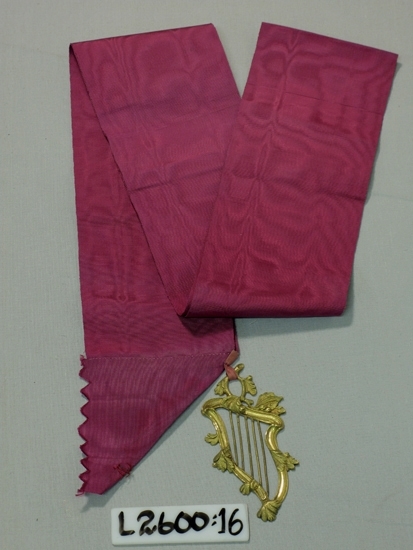 Ett 100 mm brett vinrött/cerise ripsband med vidhängande 
fantasilyra (metall).

Inskrivet i huvudboken 1933
Funktion: Ordenstecken