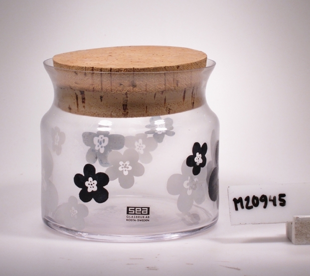 Burk med lock i kork.
Cylindrisk burk med ett avsmalnande avslut innan den utvidgande mynningen.
Ofärgat glas med målade blommor i vitt och svart.
Funktion: Burk med lock