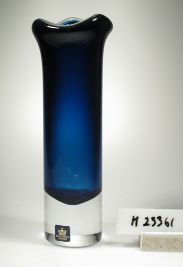 Vas. Tillverkad av Åseda glasbruk åt Kronabruken. 
Smal, cylindrisk kropp. Utsvängd, trekantig mynning och tjockt gods.
Planslipad botten. Tjock botten av ofärgat glas, resten mörkblå.
Fyrkantig, svart etikett med en krona och guldtext. Se "Signering, märkning" ovan.
Inskrivet i huvudkatalogen 1975.

Ur "Glas 75" utställningsglas.
Funktion: Vas