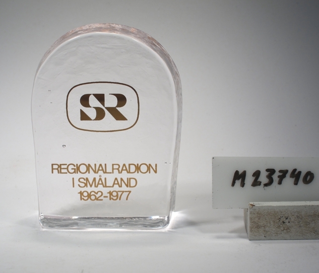 Minnesföremål från Regionalradion i Småland.
Beskrivning: Rektangulär botten, närmast lodräta väggar i nederdelen, halvcirkelformad överdel. Tryck med guldtext: "SR Regionalradion i Småland 1962-1977". (Se "Signering, märkning" 1).
Färg: Ofärgat klarglas, guldtext.
Märkning: Etikett med text och svensk flagga.
Inskrivet i huvudkatalogen 1976.

Överlämnat till regionalradions tjänstemän m.fl. i samband med avslutningsfesten, Evedal 1977-08-26.
Funktion: Minnesföremål