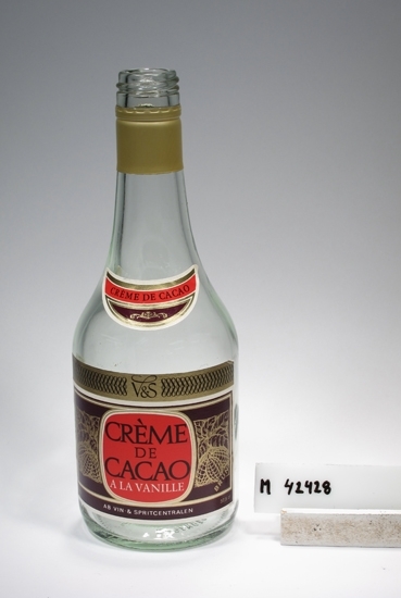 Likörflaska " Crème de cacao", 37,5 cl.
Beskrivning: Låg, cylindrisk kropp. Rundad övergång, insvängd - konisk, stor hals med gänga.
Etikett fram: Halmånformad etikett i vitt och rött, brunt och guld med text: " Créme de cacao"
Etikett bak: Stor rektangulär etikett i samma färger, med text: " V&S, Créme de cacao a la vanille: 24% A.B Vin & Spritcentralen 37,5 cl"
Färg: Ofärgat klarglas.
Relief i botten: Engångsglas, återtages ej. PLM. H 38 (?) 37,5 cl.
I Huvudkatalogen anges måttet 3,7 cl, vilket ej stämmer.
Inskrivet i huvudkatalogen tidigast 1983.
Funktion: Likörflaska