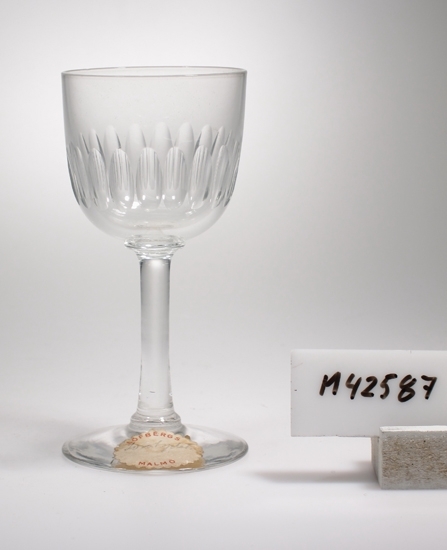 Brännvinsglas, med täta lodräta olivskär (linjer) 
på den övre delen av kupan.
Rakt ben och fot.
Klarglas.
Oval tandad lapp :" Löfbergs Malmö" (och oläslig text).

Se jub.kat. 1871-1931 sid. 18.
Enl. reg.kort. " Ulle" med täta oliver endast i denna katalog, 
i några andra har den skär med hack (slipskär).

Gåva av Sten Löfberg 1984.
Funktion: Brännvinsglas