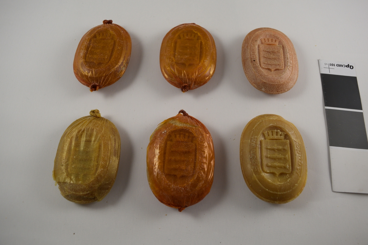 Oval såpe, seks stykker som er tilnærmet like. Produsert ved Saanums Sepefabrikk i Mandal. Tre stykker pakket i oransje plastikk (a-c), en i brun plastikk (d). To uten plastemballasje (e-f), men i ulik farge. Alle dekorert med Mandals kommunevåpen, med tre laks.