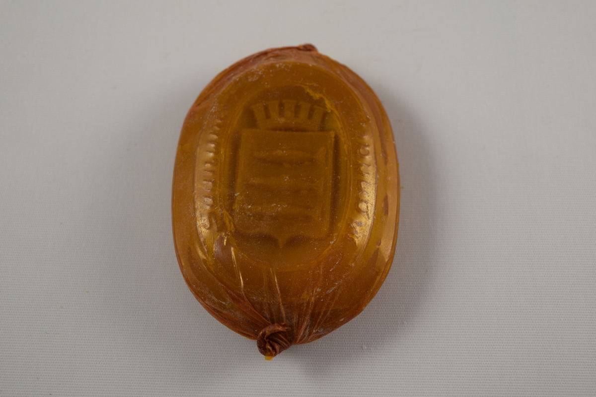 Oval såpe, seks stykker som er tilnærmet like. Produsert ved Saanums Sepefabrikk i Mandal. Tre stykker pakket i oransje plastikk (a-c), en i brun plastikk (d). To uten plastemballasje (e-f), men i ulik farge. Alle dekorert med Mandals kommunevåpen, med tre laks.
