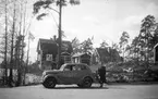 Bostadshus, kvinna vid en bil, en Ford Eifel 1939, och ett Café.