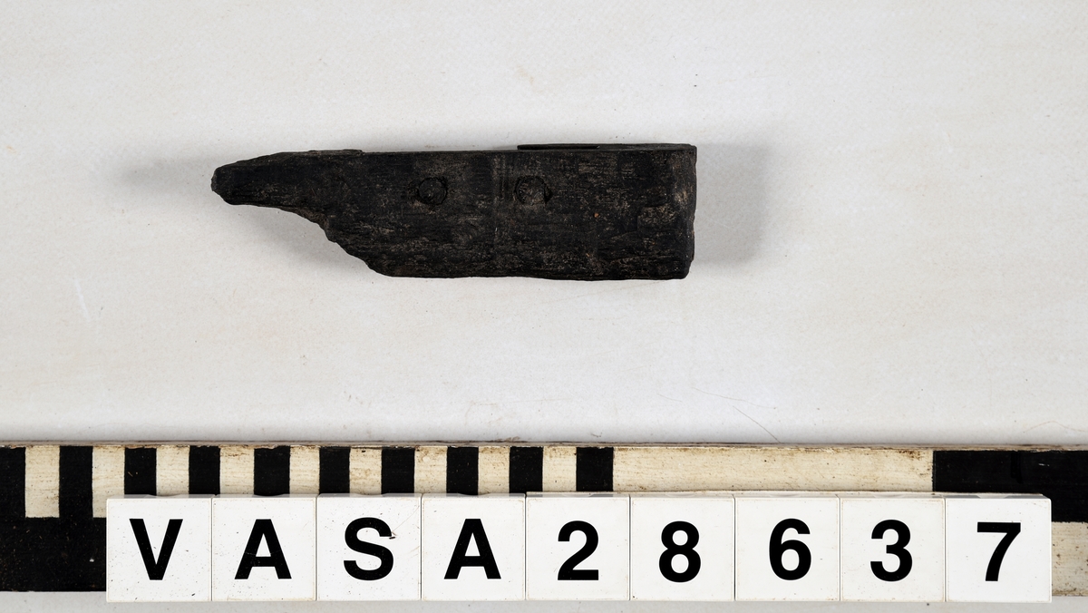 Avbruten del av ram, med rektängulärt urtag för att sammanfoga föremålet med ett annat föremål. Två pluggar är kvar i urtaget.  Nästan halva föremålet avsplittrat. Insidan på urtaget välbevarat med räta vinklar.