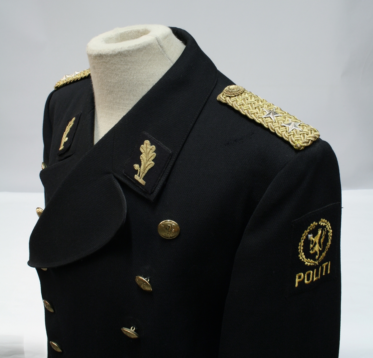 Uniform for politinspektør etter 1963-reglement med merking etter 1977-reglement. Helårsuniform M/1963 med skjorte, langbukse, belte, helårskappe og jakke. Skyggelue med gullbrodert bånd, loslue og båtlue M/1967. Kappen og jakken har skulderklaffer av flettede gullsnorer med to sølvstjerner og festeknapp i gullbroderi. Skjorten har en enklere skulderklaff med to gule render og to sølvstjerner. Det følger med et ekstra enkelt lærbelte og et ekstra sett skulderklaffer til skjorte. Plaggene har gullfargede løveknapper.