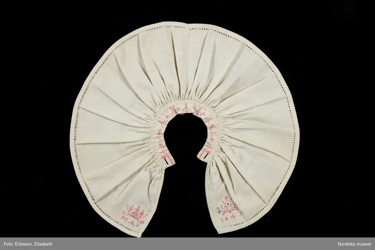 a. Kvinnokrage i form av ett rynkat krås, av halvlinnelärft med rosa korssömsbroderi, märkt MAD 1847
b. Tillhörande länkknapp av mässing.
/Berit Eldvik 2010-09-21