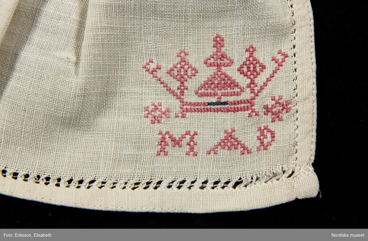 a. Kvinnokrage i form av ett rynkat krås, av halvlinnelärft med rosa korssömsbroderi, märkt MAD 1847
b. Tillhörande länkknapp av mässing.
/Berit Eldvik 2010-09-21