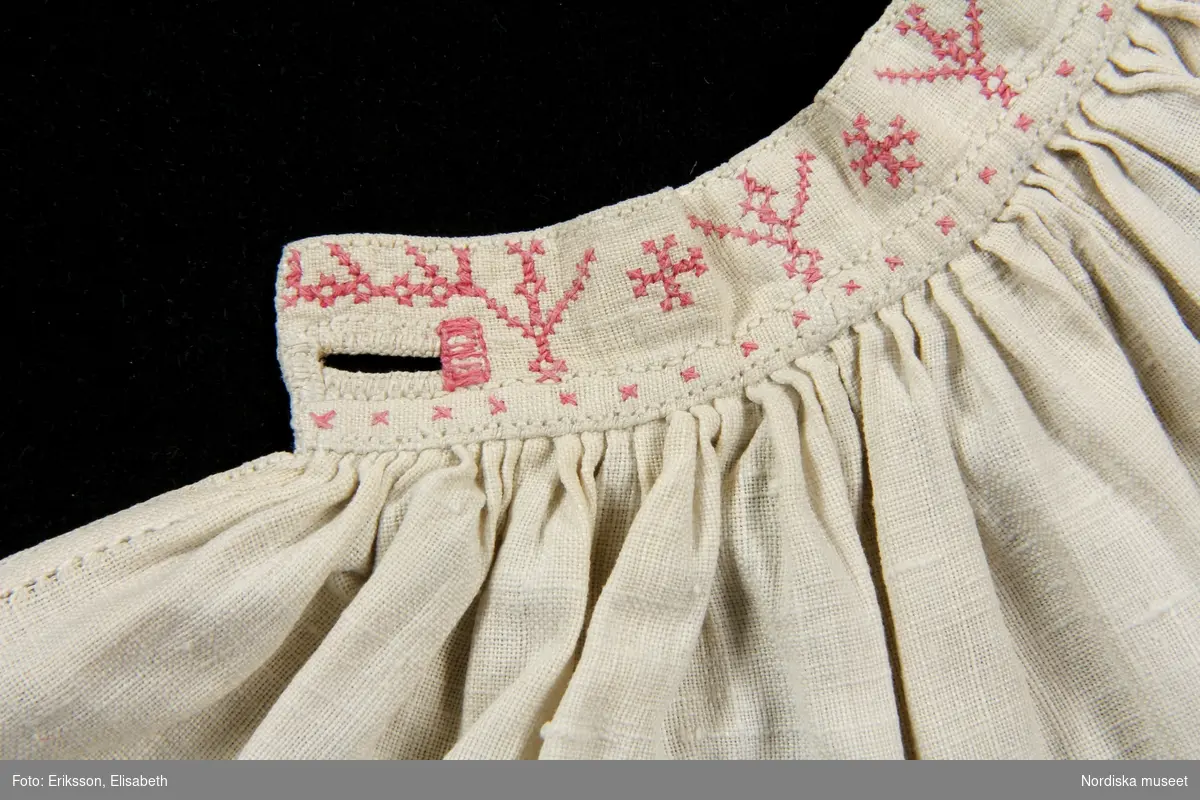 a. Kvinnokrage i form av ett rynkat krås, av halvlinnelärft med rosa korssömsbroderi, märkt MAD 1847
b. Tillhörande länkknapp av mässing.
/Berit Eldvik 2010-09-21