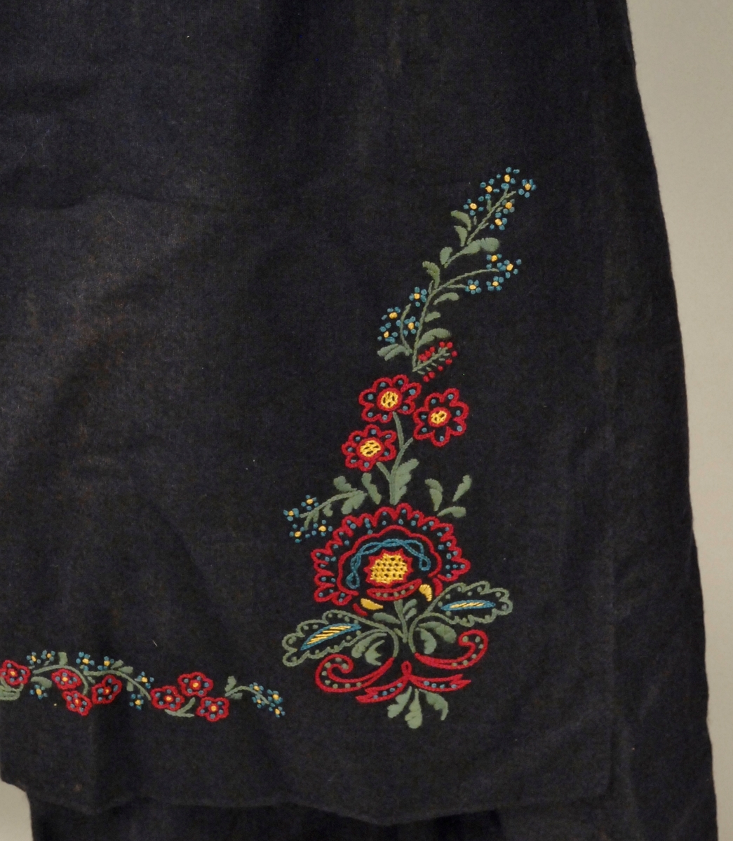 Ullstoff i blått. Drakt og forkle. Foret livstykke. Brodert mønster foran på livstykket og nederst foran på forkle, øverst på ryggstykke og rundt halsen. Forkle knyttet sammen med silkebånd.