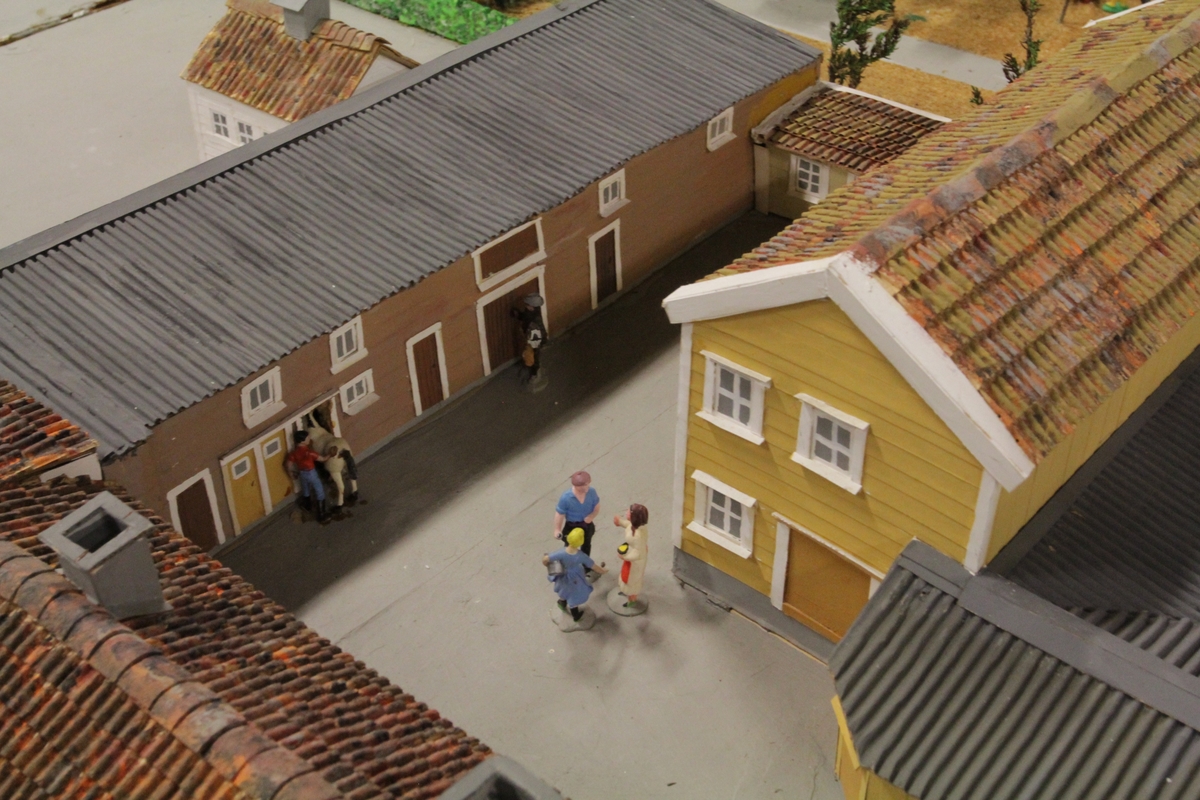 Stor rektangulær modell bygget av tremateriale og tilført mindre gjenstander av annet materiale for å gjøre bymodellen så levende og autentisk som mulig.Beplantning, kjøretøy, mennesker og aktivitet i bakgårdene.  Den gamle lærerskolen som midtpunkt - og gjerdes inne av Elvegaten, Tollbodgaten, Rådhusgaten og Kronprinsens gate i Kristiansand. Laget i målestokk 1:50