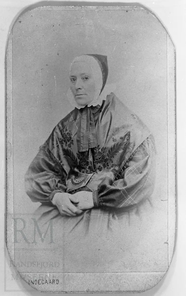 Portrett av Kari Olsdatter Hoff , gift Kari Haugtvedt (1822-1898), sittende i rutet kjole, honnlue med skru og med silkesjal over skuldrene.