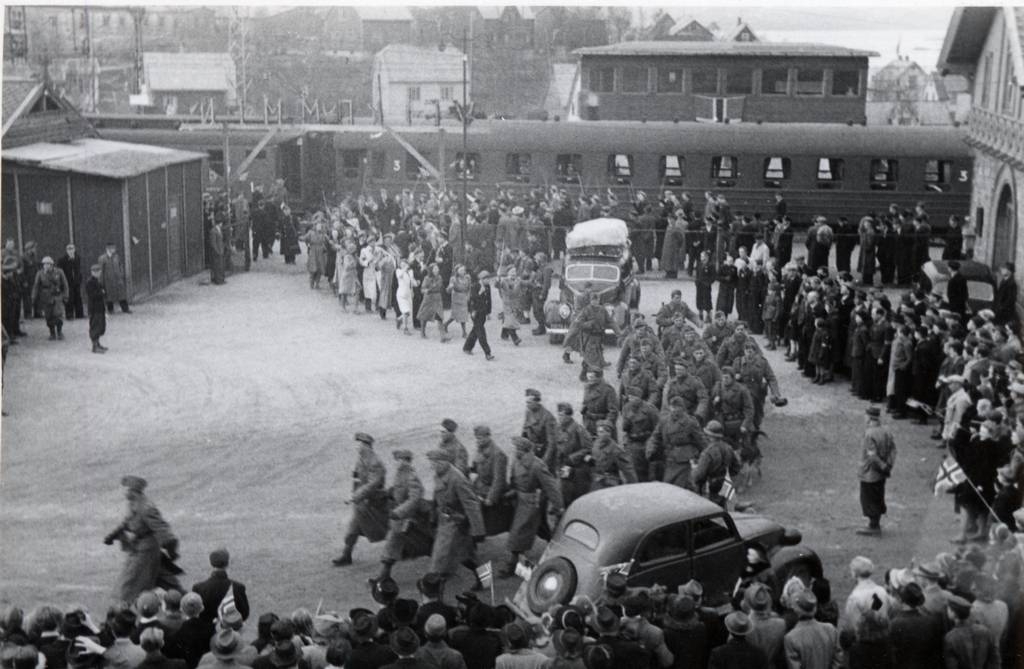 De norske soldatene marsjerer ut fra Narvik stasjon. Som baktropp kom Russen Anno 1945.Ved plattforme en personvogn, 3. klasse. Bak vognen sees stilverket på Narvik stasjon.