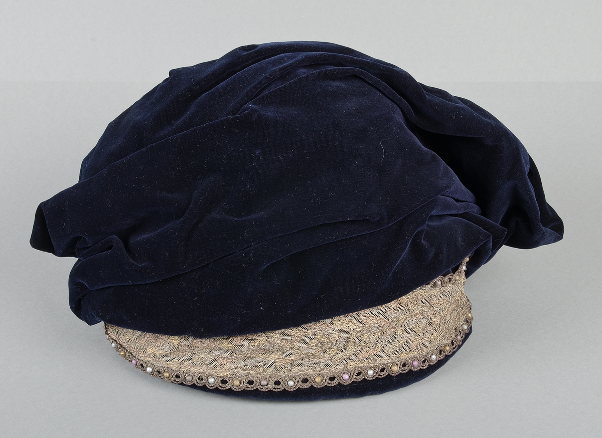 Hatt av blå sammet, prydd med tyllspets med guld och silvertrådar samt pärlor i guld silver och lila. Fodrad med svart siden. I botten med guldtryck: Alma Engelbreckt Modeaffär, Gamla Torget 2, Uppsala. Svart sammetsvalk i kullen.
