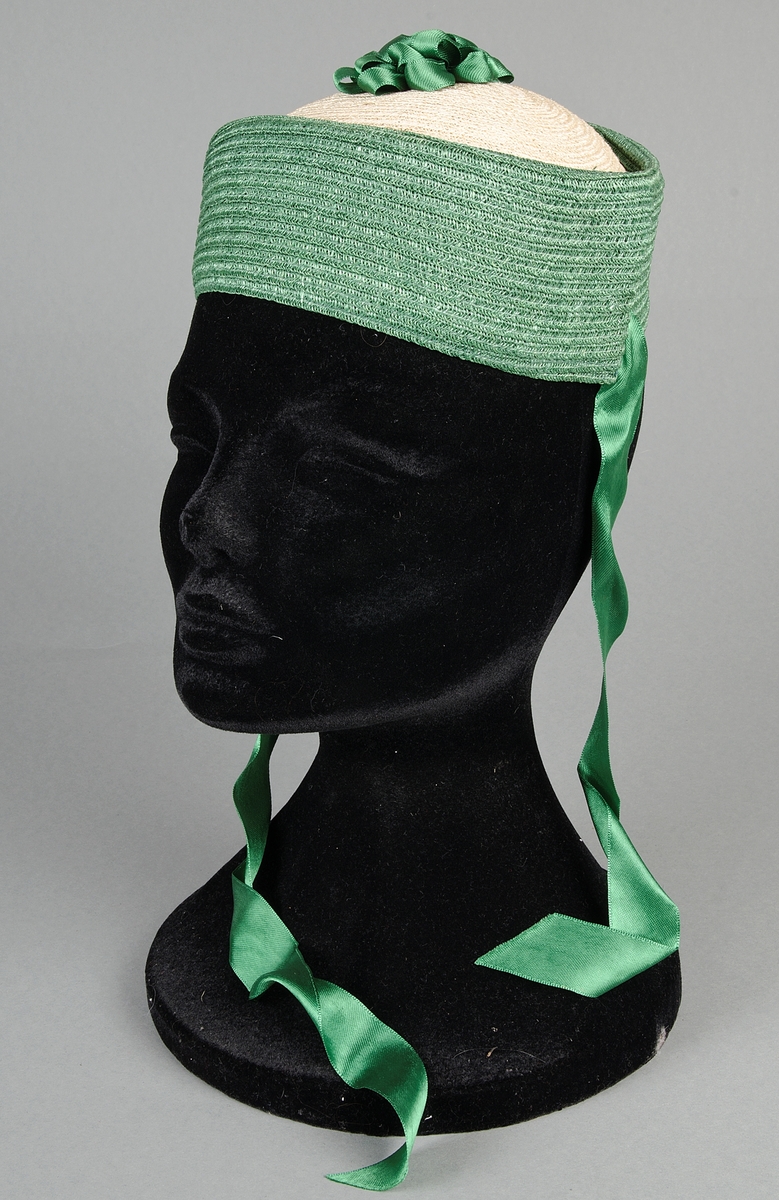 Damhatt av flätad strå i vitt och grönt. Rosett och band (50 cm långa) av grönt siden. Invändigt kantad med grönt spencerband. Brun prislapp. Storlek 51. Pris 6.50.