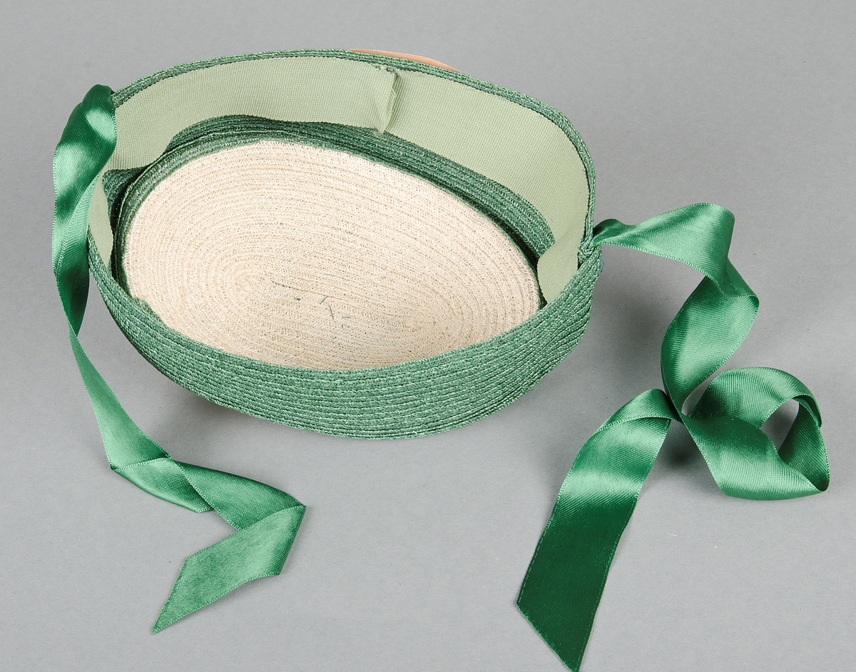 Damhatt av flätad strå i vitt och grönt. Rosett och band (50 cm långa) av grönt siden. Invändigt kantad med grönt spencerband. Brun prislapp. Storlek 51. Pris 6.50.