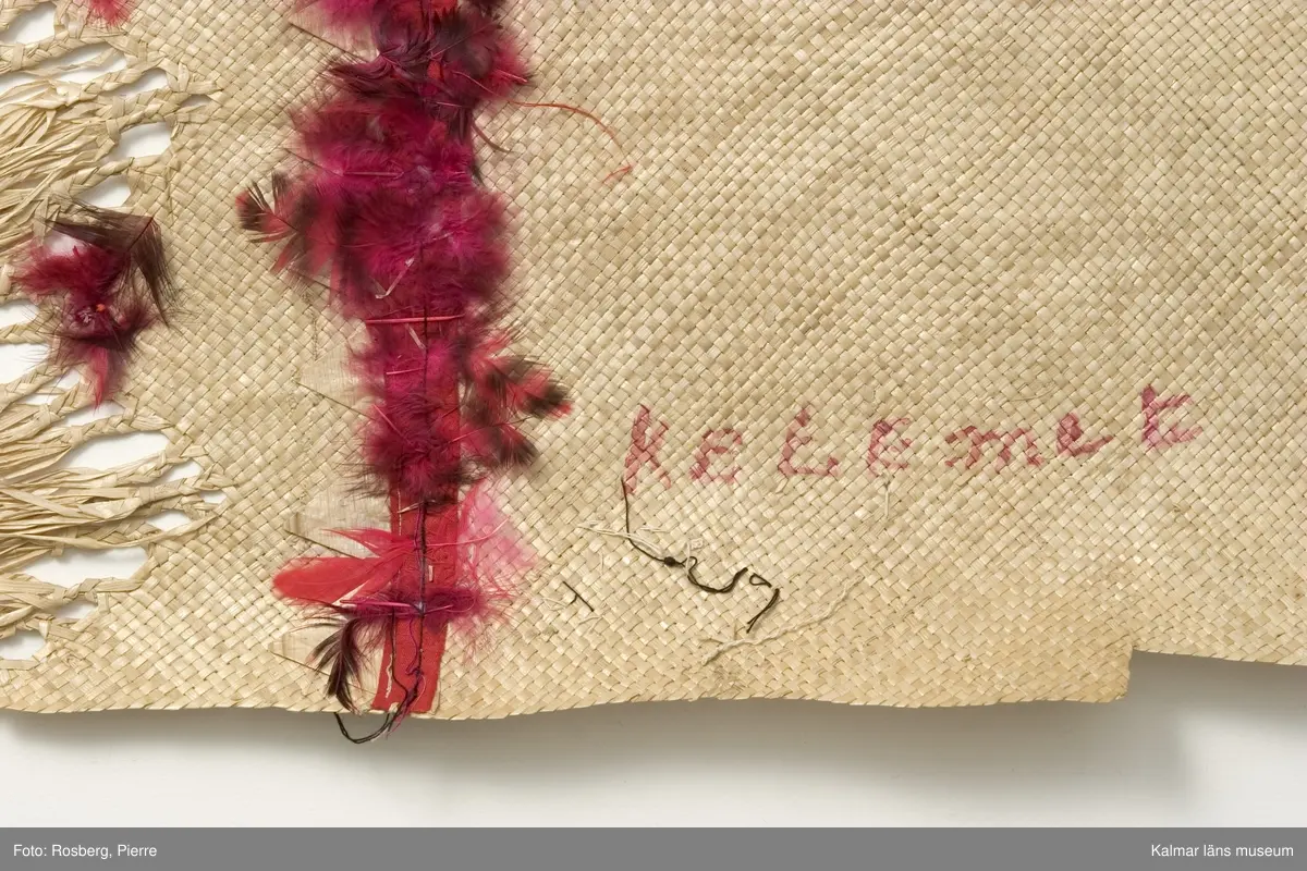 KLM 28465. Matta/kjol av pandanus (en palmväxt) och fjädrar. Plagget benämns 'ie toga. 'ie är en av tre sorters odlade palmväxter som används av samoanerna för mattorna. Mattan är mer att betrakta som ett ceremoniellt klädesplagg flätat av pandanus med fransar i båda ändarna och dekorerat med röda och några få vita fjädrar fastsydda med maskinsöm. Den här mattan, är ett ceremoniellt klädesplagg, den bärs runt midjan som en kjol, dubbelvikt så att man skall kunna se fransarna. Den hålls vanligen på plats av en gördel av barktyg. Följande namn finns påskrivna, på framsidan, KELEmet, TutuilA x Elsa, LUIT; på baksidan, AiLUA Poliko, LEALA SOLA. Därutöver ett nummer som tycks vara 10 finns också. Tutuila är för övrigt namnet på en sydöstsamoansk ö, vilken är huvudön i Amerikanska Samoa.
