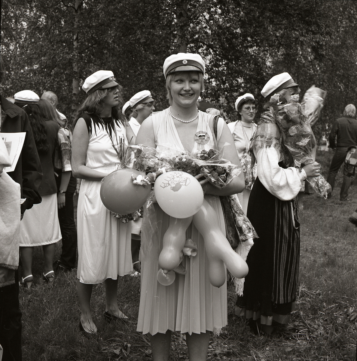 En kvinna poserar i studentmössa på Ohnbacken i Bollnäs 1981. Hon bär en ljus klänning och håller i i famnen blommor, ballonger och presenter. Bakom kvinnan myllrar av studenter och deras familjer som gratulerar med blommor på examensdagen.