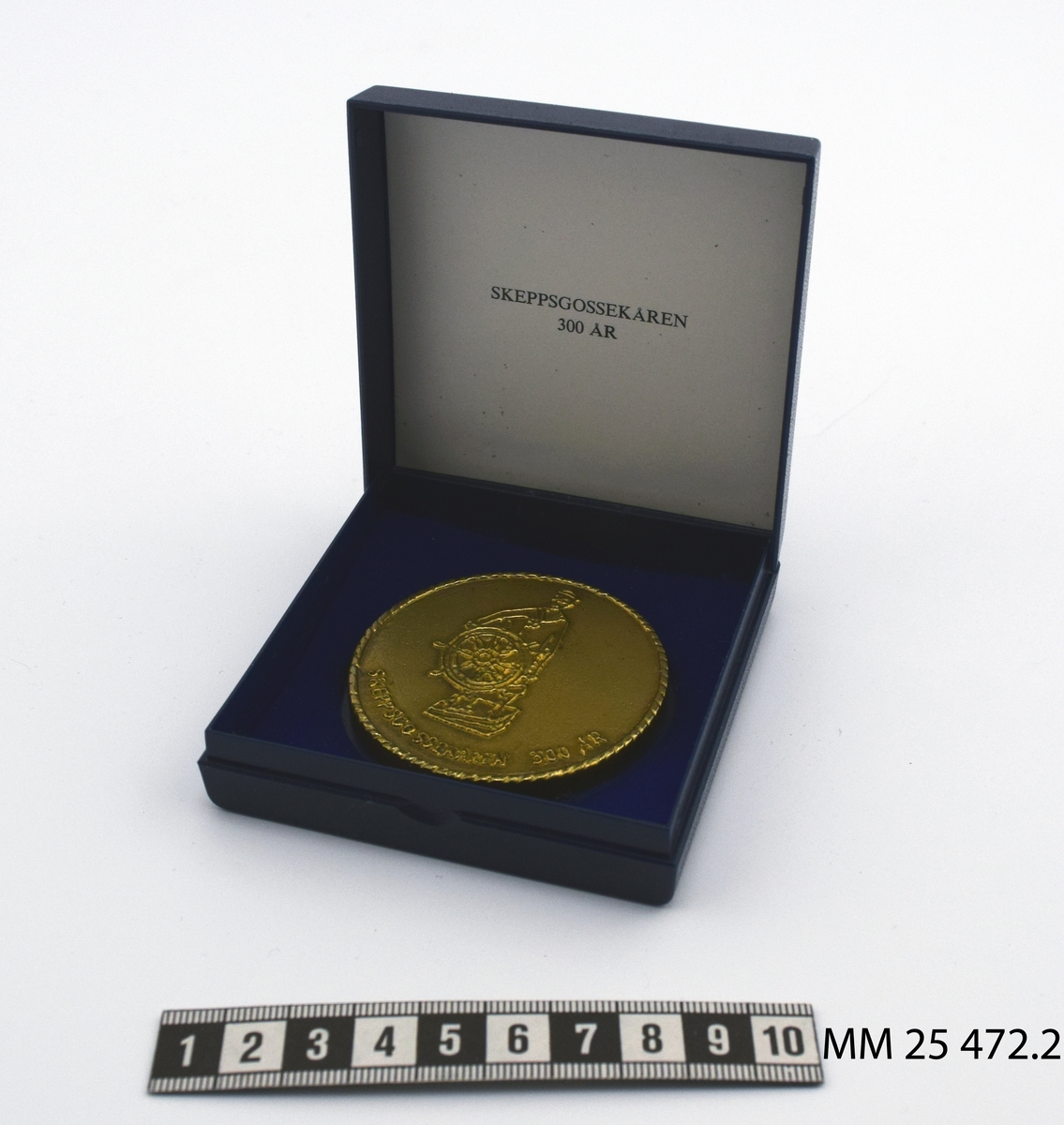 Rund plakett av gulmetall med präglat motiv. På ena sidan motiv av skeppsgosse vid styrratt samt texten: Skeppsgossekåren 300 år". På andra sidan motiv av korvetten Carlskrona samt texten: "Karlskrona 1685-1985" "Korvetten Karlskrona 1841". Plaketten förvaras i blå plastask med sammetsklädd insida.