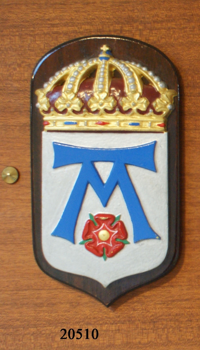 Vapensköld gjuten av mässing. Krönt med kunglig krona, målad i heraldiska färger: guld, silver blått och rött. Bottnen i silver är försedd med emblem i blått format format som ett M med en balk över. Nedanför emblemet finns en röd blomma med fem kronblad. Guld i mitten och gröna småblad mellan kronbladen.

Skölden är fäst till en fernissad platta av mörk mahogny.