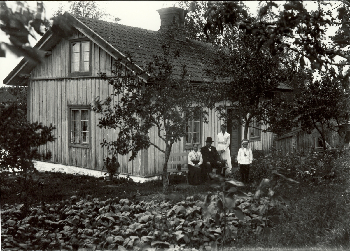 En familj avporträtterad utanför en stuga i Floda, Hjorted, 1919. Petter Wilhelm Pettersson f 1856 (slöjdare av likkistor m.m.), hustrun Anna Lovisa Nilsdotter f 1848, deras dotter Alma Elisabet f 1886 samt Almas son Gustaf Einar (Åsberg) f 1910.