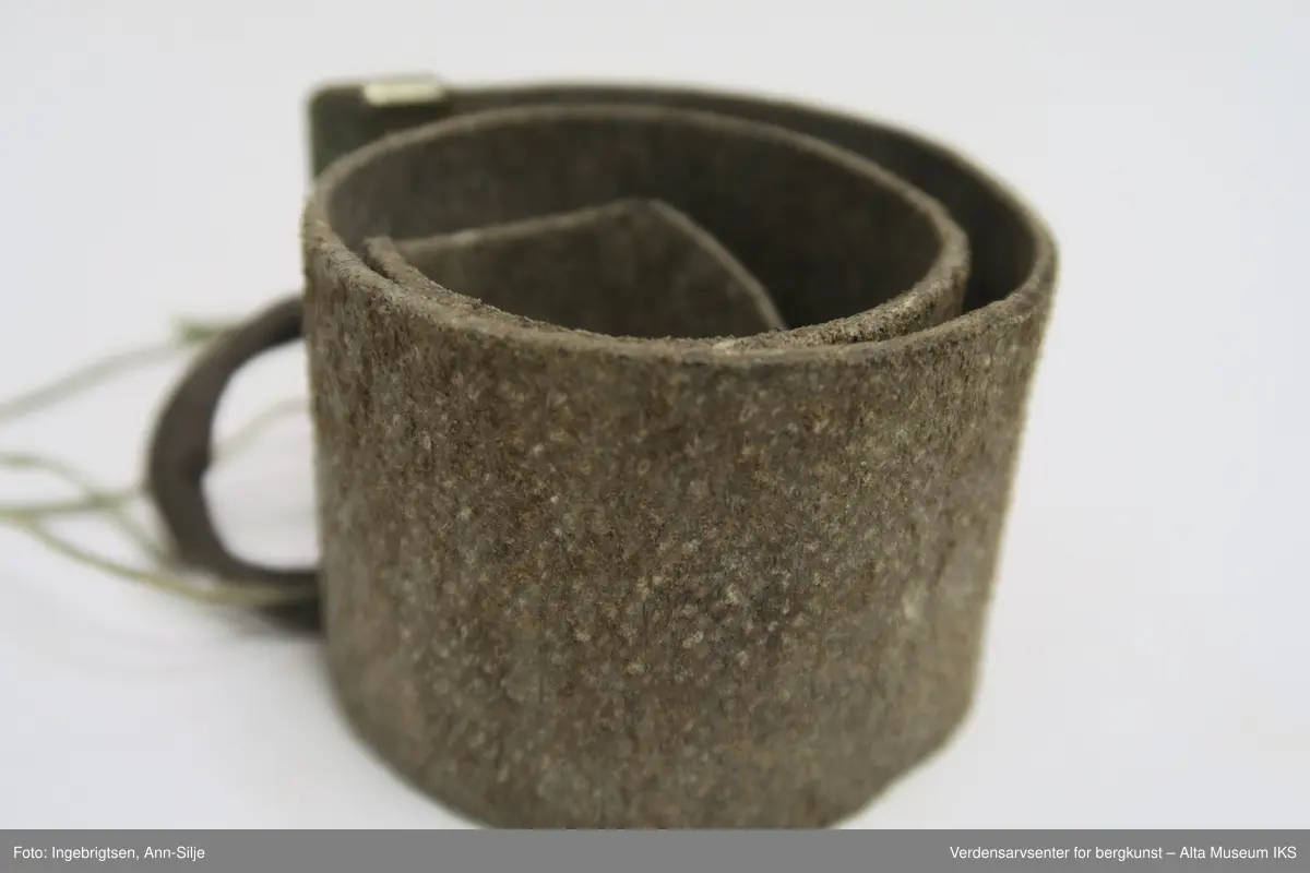 En rektangulær lærremse med et metallstykke klemt i den rette enden. Remsen ender i en spiss i den andre enden. En løkke av tau er festet i metallet. På oversiden av læret er det rutemønster i læret, mens undersiden er glatt.