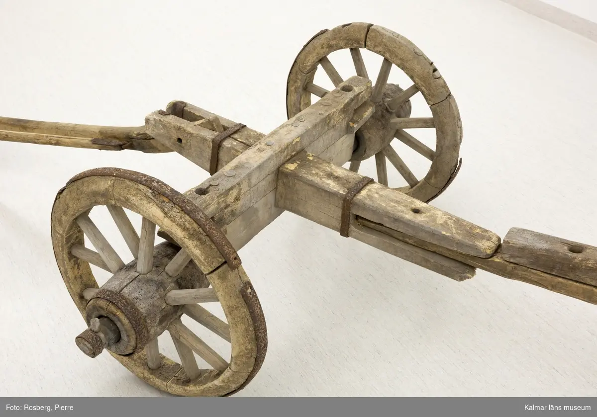 KLM 11833. Bultvagn, hästvagn. Av trä och järn. För hästar. Bultvagn som består av två hjulpar med två bankar, fyra träaxlahjul med järnbeslag, skakel samt dräkt. Det främre hjulparet är mindre än det bakre och har en tillhörande skakel som fästes i en träanordning på dess bank. Dräkten består av en kraftig rektangulär stång av trä med sex stycken borrade hål. Dräkten fästes i en annan träanordning som sitter på motsatt sida av skakeln på det främre hjulparets bank. På det bakre hjulparets bank sitter ett bågformat hål där dräkten sedan förs igenom för att sammankoppla vagnens delar samt justera dess längd. De bakre hjulens nav är dekorerade med zig-zag mönster.