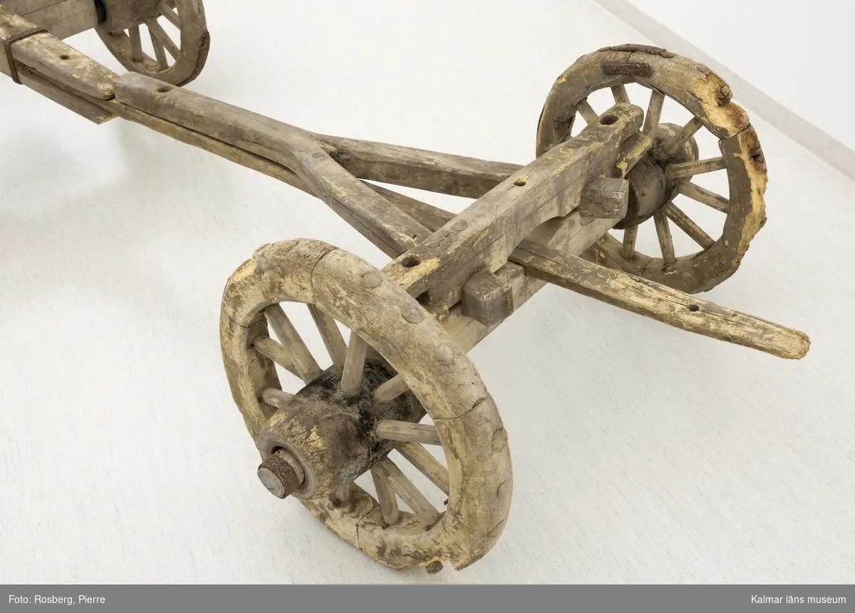 KLM 11833. Bultvagn, hästvagn. Av trä och järn. För hästar. Bultvagn som består av två hjulpar med två bankar, fyra träaxlahjul med järnbeslag, skakel samt dräkt. Det främre hjulparet är mindre än det bakre och har en tillhörande skakel som fästes i en träanordning på dess bank. Dräkten består av en kraftig rektangulär stång av trä med sex stycken borrade hål. Dräkten fästes i en annan träanordning som sitter på motsatt sida av skakeln på det främre hjulparets bank. På det bakre hjulparets bank sitter ett bågformat hål där dräkten sedan förs igenom för att sammankoppla vagnens delar samt justera dess längd. De bakre hjulens nav är dekorerade med zig-zag mönster.