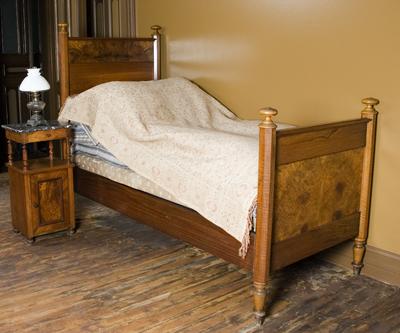 En oppredd enkeltseng i tre står i et gulmalt rom, med et nattbord med en lampe ved siden av. 