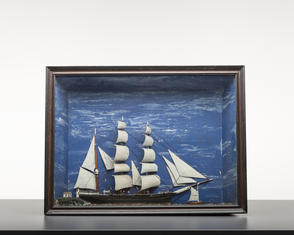 Skutemodell i "trapesformet" skrånende kasse. Til å henge på veggen. Skuten er tremastet. Det er også  også er et fyr med hus og en liten båt med 2 seil i kassen. Kassen skråner innover mot vegg.