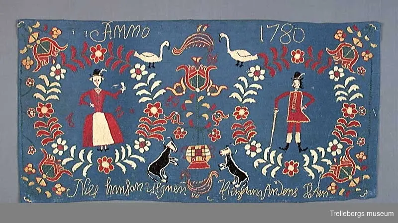 På en mellanblå botten är två kransar broderade, i den ena kransen står en kvinna och i den andra en man. I dynans hörn är blommor broderade, samma blomma kommer igen i dynans mitt tillsammans med en tupp, två gäss och två kor. Broderiets färger är rött, vitt, gult, grönt och svart. Dynan är märkt Anno 1780 Nils Hansson Ulgren Kierstin Andersdotter.