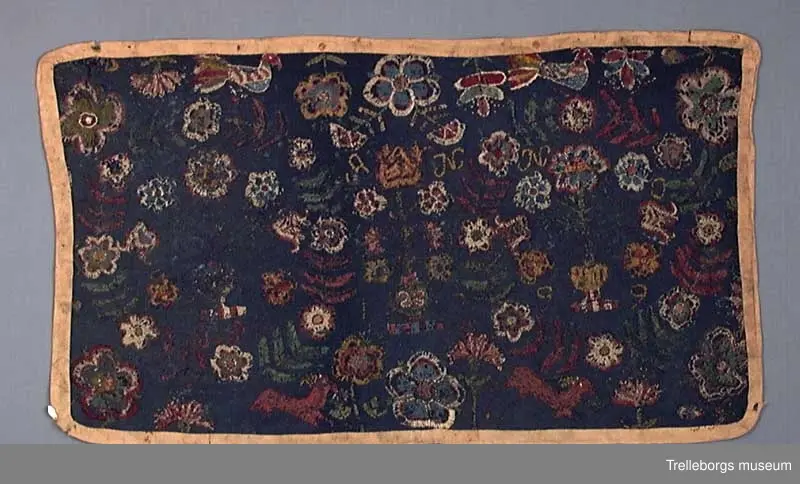 Ylletyg i tuskaft i botten, broderi 1 tr ullgarn.
Dyna av sydd flossa på blå vadmal röd kant, mönster, flera färger. Plattsöm. Anno 1799 från Gärdslöv. 
Broderiet är mkt slitet, men 2 stora kransar av blad och blommor med en blomstervas med 7 blommor i kan tydligt urskönjas. Ovanför vardera kransen finns en fågel. 
Mittfigur är en blomstervas med blommor.
Nedtill på agedynan hoppar 2 st hjortar.
Färger: mörkrött, rödlila, vitt, grönt, gult och ljusblått.
Dynan är kantad med brunt bomullstyg.
Agedynan är från Gärdslöv. 
Växter och frukter. Enligt Ernst Fischer, Årsbok från Malmö museum 1971: "Nästa åkedyna tillhör Tbgs. museum. Även den överensstämmer till alla delar med gruppens andra dynor med det undantaget, att blommorna i vasen inom kransarna är mera naturalistiskt utformade och därigenom närmar sig ursprungsdynan från 1713. Däremot är vasen mellan kransarna mer lik den föregående åkdynan.
Broderiet som är märkt ANNO 1799 och en signatur, av vilken man kan tyda IP, kommer från Gärdslöv, Vemmenhögs hd. Den bygger på samma förlaga som de övriga inom gruppen, men brodösen har icke haft samma skicklighet som de, vilka utfört dessa. Hon har förenklat teckningen och inte nyanserat detaljerna i samma utsträckning."
Sid 144-145.