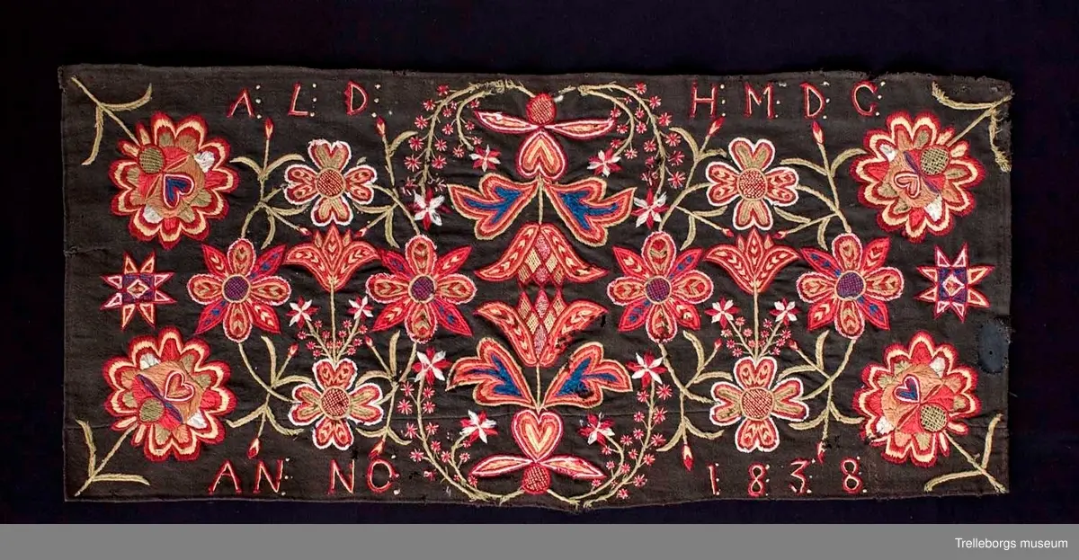 Botten är svartbrun tuskaft i ullgarn. 
Ringkrans med 4 blommor.
Hela mönstret är spegelvänt utmed en längsgående axel (frånsett 2 tulpanfigurer och märkningen ALD HMDG utmed den övre långsidan och ANNO 1838 utmed den nedre långsidan. 
Broderiet består av gängse fantasiblommor och tulpaner, omgivna av bladslingor.
Färgerna är vitt, blekgult, blekgrönt, blått, bronsgult mm