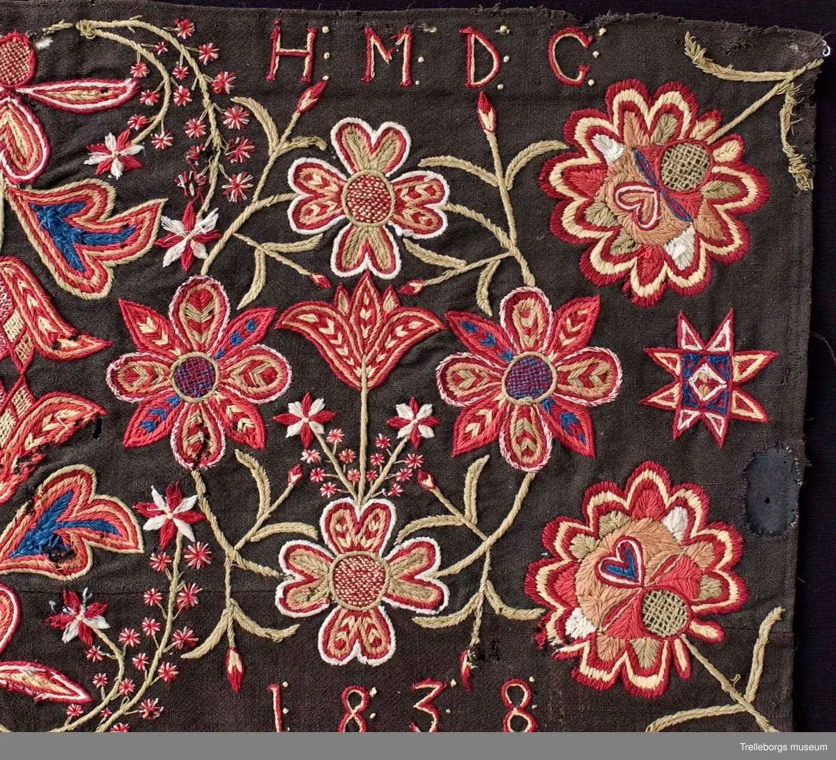 Botten är svartbrun tuskaft i ullgarn. 
Ringkrans med 4 blommor.
Hela mönstret är spegelvänt utmed en längsgående axel (frånsett 2 tulpanfigurer och märkningen ALD HMDG utmed den övre långsidan och ANNO 1838 utmed den nedre långsidan. 
Broderiet består av gängse fantasiblommor och tulpaner, omgivna av bladslingor.
Färgerna är vitt, blekgult, blekgrönt, blått, bronsgult mm