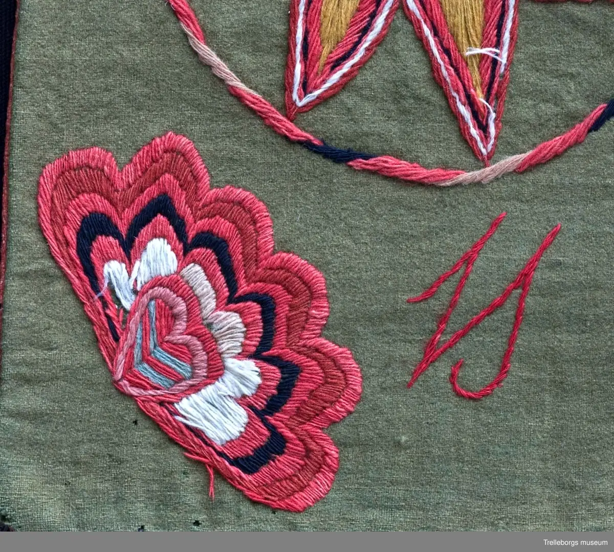 Bottentyg är grön vadmal. Tre stora ringar i kontur utgör huvudmotiv. I ringen i mitten är en stor blomma med 8 kronblad utfyllnad. I de båda ringarna vid sidan är broderat en spetsuddig stjärna med 8 vingpar. Dess centrum är en snedställd kvadrat, sydd i gallerliknande konstsöm. Utmed agedynans långsidor är broderat olika blomsterstjärnor, pionliknande blommor och en blomliknande rundning av 4 st hjärtfigurer. Färger: vitt, gult, svart, blågrått samt många olika nyanser i rött. Baksidan tegelröd yllevävnad. 
Enligt katalogen märkt I.W.D.S.  SL. Hjärtrosetter och hjärtan. 
Enligt Ernst Fischer, Årsbok från Malmö museum 1971, sid 180: En åkdyna i Tbgs museum har som mittmotiv en runduddig stjärna, omgiven av en ring samt en hjärtrosett över och under, flankerad av spetsuddiga stjärnor. T.h. och v om mittmotivet stora spetsuddiga stjärnor inom ringar. I hörnen varierande blomformer. De ganska svårtydda initialerna skulle kunna tydas som LWD IVH, ehuru bokstäverna då icke är placerade i rätt ordning. IVH skall i så fall beteckna I Vämmenhög.