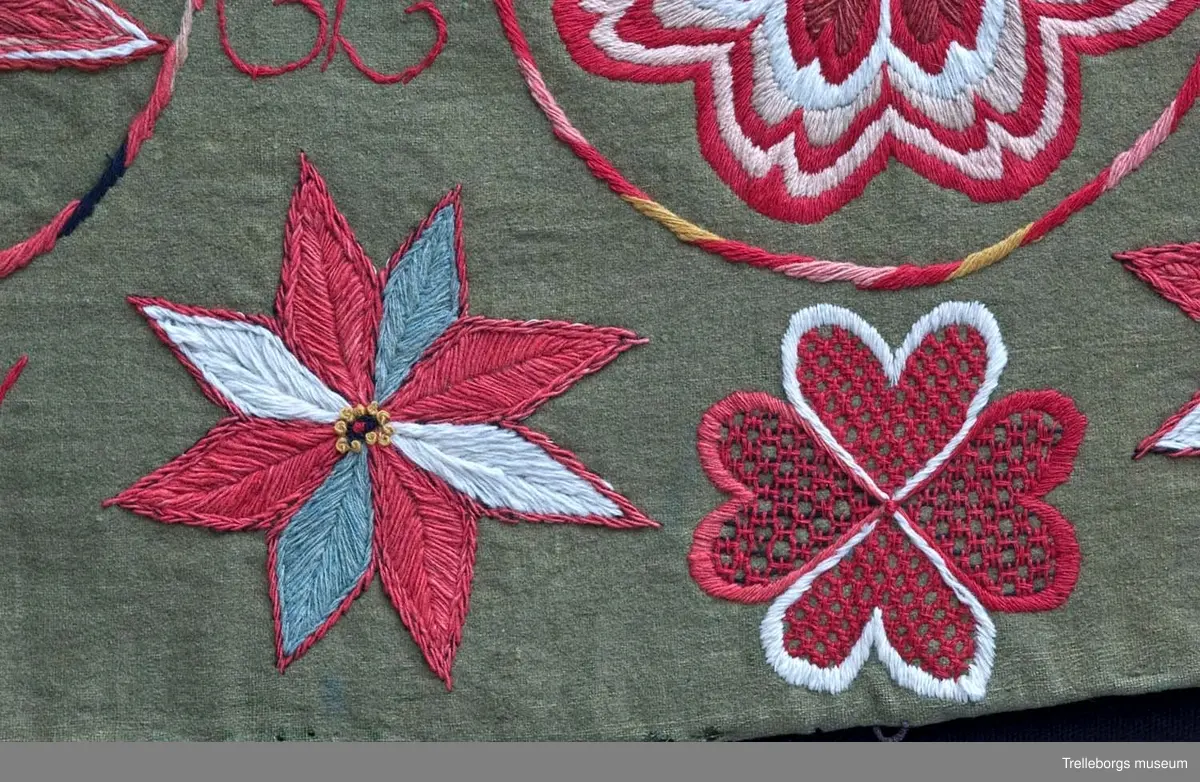 Bottentyg är grön vadmal. Tre stora ringar i kontur utgör huvudmotiv. I ringen i mitten är en stor blomma med 8 kronblad utfyllnad. I de båda ringarna vid sidan är broderat en spetsuddig stjärna med 8 vingpar. Dess centrum är en snedställd kvadrat, sydd i gallerliknande konstsöm. Utmed agedynans långsidor är broderat olika blomsterstjärnor, pionliknande blommor och en blomliknande rundning av 4 st hjärtfigurer. Färger: vitt, gult, svart, blågrått samt många olika nyanser i rött. Baksidan tegelröd yllevävnad. 
Enligt katalogen märkt I.W.D.S.  SL. Hjärtrosetter och hjärtan. 
Enligt Ernst Fischer, Årsbok från Malmö museum 1971, sid 180: En åkdyna i Tbgs museum har som mittmotiv en runduddig stjärna, omgiven av en ring samt en hjärtrosett över och under, flankerad av spetsuddiga stjärnor. T.h. och v om mittmotivet stora spetsuddiga stjärnor inom ringar. I hörnen varierande blomformer. De ganska svårtydda initialerna skulle kunna tydas som LWD IVH, ehuru bokstäverna då icke är placerade i rätt ordning. IVH skall i så fall beteckna I Vämmenhög.