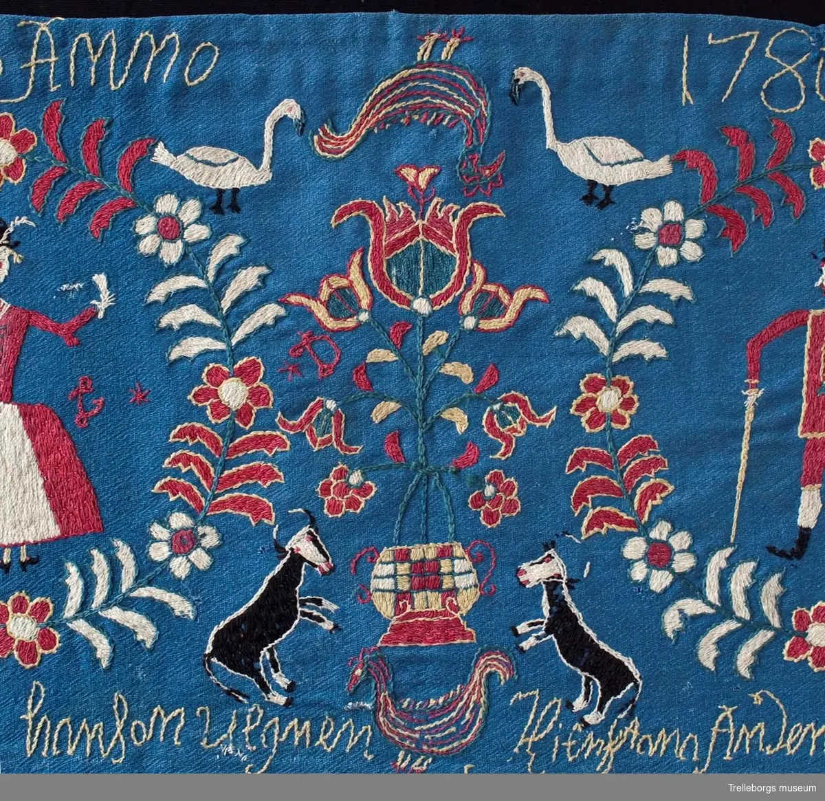 På en mellanblå botten är två kransar broderade, i den ena kransen står en kvinna och i den andra en man. I dynans hörn är blommor broderade, samma blomma kommer igen i dynans mitt tillsammans med en tupp, två gäss och två kor. Broderiets färger är rött, vitt, gult, grönt och svart. Dynan är märkt Anno 1780 Nils Hansson Ulgren Kierstin Andersdotter.