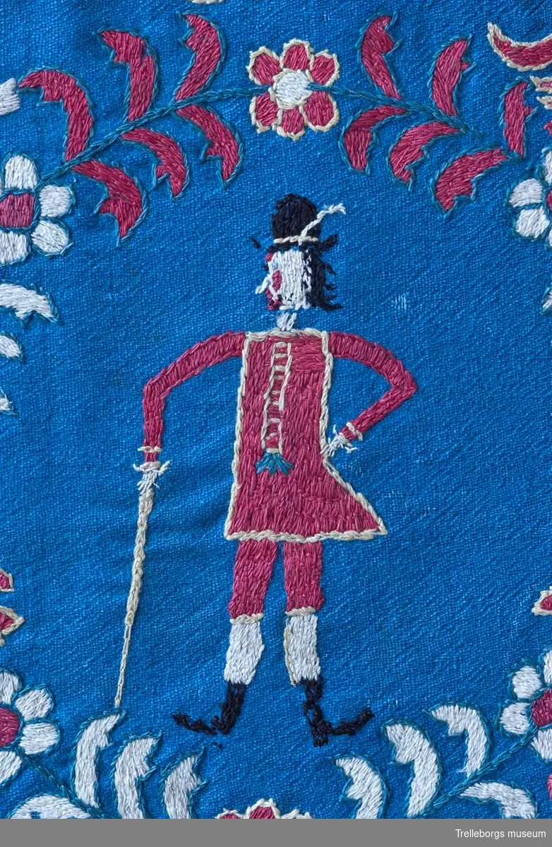 På en mellanblå botten är två kransar broderade, i den ena kransen står en kvinna och i den andra en man. I dynans hörn är blommor broderade, samma blomma kommer igen i dynans mitt tillsammans med en tupp, två gäss och två kor. Broderiets färger är rött, vitt, gult, grönt och svart. Dynan är märkt Anno 1780 Nils Hansson Ulgren Kierstin Andersdotter.