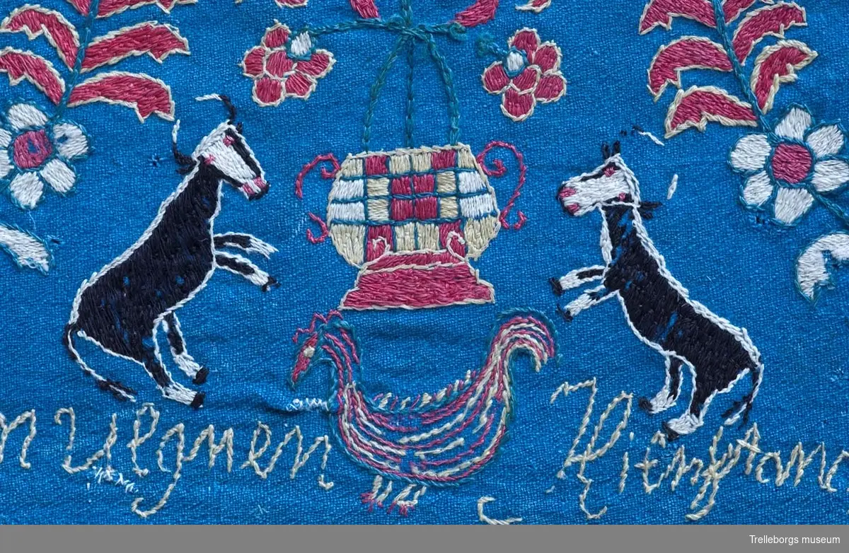 På en mellanblå botten är två kransar broderade, i den ena kransen står en kvinna och i den andra en man. I dynans hörn är blommor broderade, samma blomma kommer igen i dynans mitt tillsammans med en tupp, två gäss och två kor. Broderiets färger är rött, vitt, gult, grönt och svart. Dynan är märkt Anno 1780 Nils Hansson Ulgren Kierstin Andersdotter.