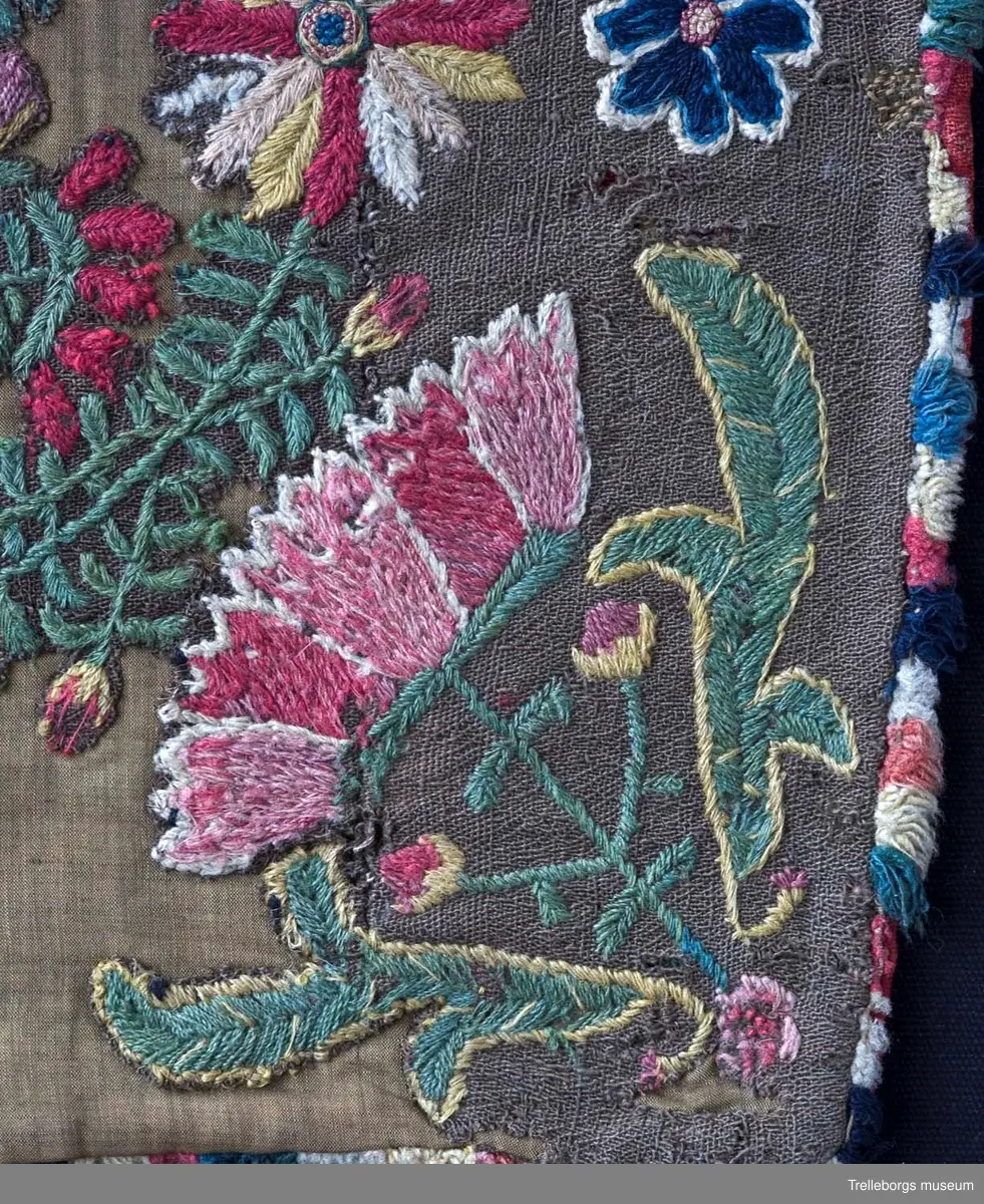 Broderad agedyna på grov, brun yllebotten. Motivet är två grönskande blomsterkransar fyllda med vad som mest liknar blommande buskar, även mellan kransarna finns två blommande buskar. Här är dynan märkt med CAD i den ena och ANO i den andra, båda på ett litet fält med franska knutar i vitt och rött. Under busken med ANO står 180 ganska glest, sista siffran finns inte kvar. Dynan är monterad med en flerfärgad kavelfrans och en klarröd, ganska grov yllebaksida.