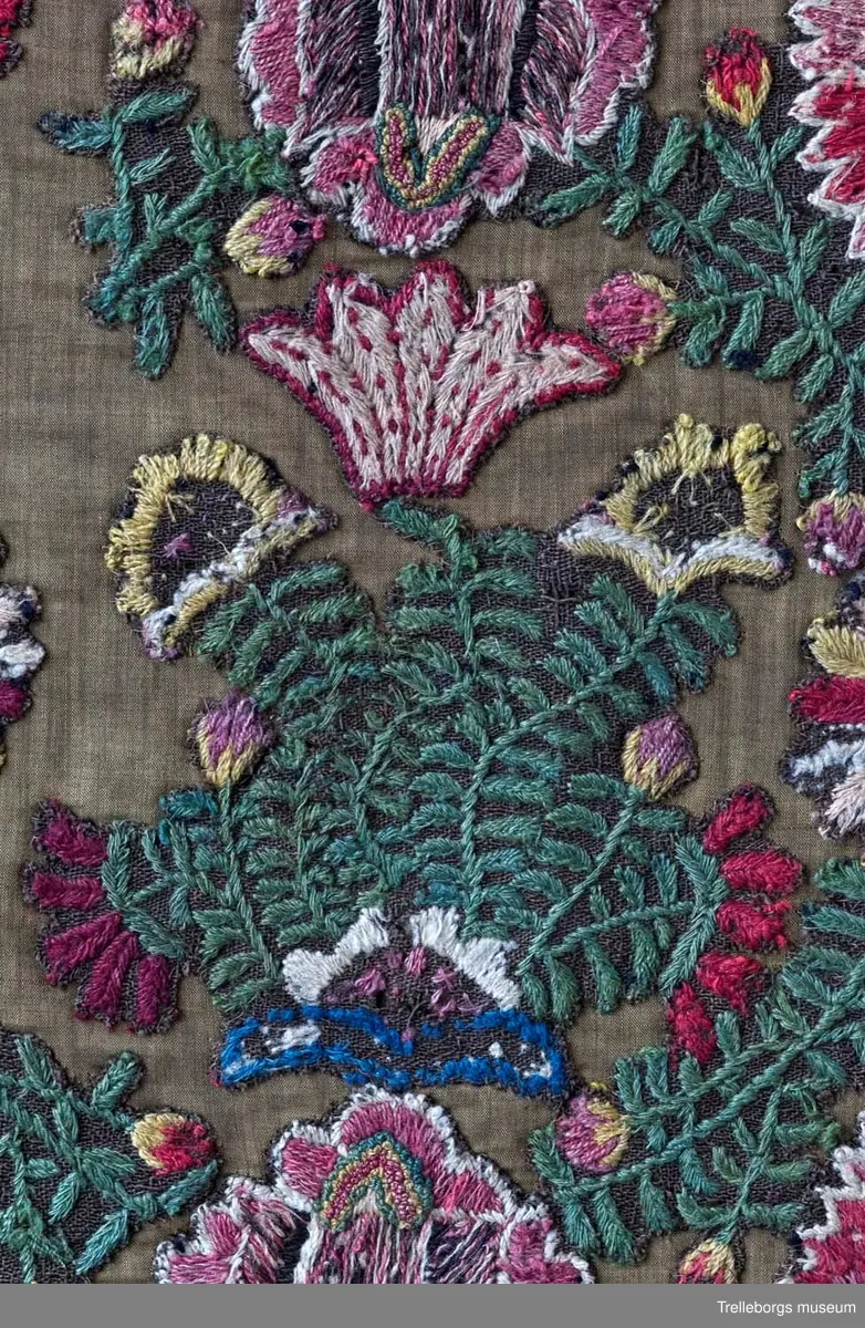 Broderad agedyna på grov, brun yllebotten. Motivet är två grönskande blomsterkransar fyllda med vad som mest liknar blommande buskar, även mellan kransarna finns två blommande buskar. Här är dynan märkt med CAD i den ena och ANO i den andra, båda på ett litet fält med franska knutar i vitt och rött. Under busken med ANO står 180 ganska glest, sista siffran finns inte kvar. Dynan är monterad med en flerfärgad kavelfrans och en klarröd, ganska grov yllebaksida.