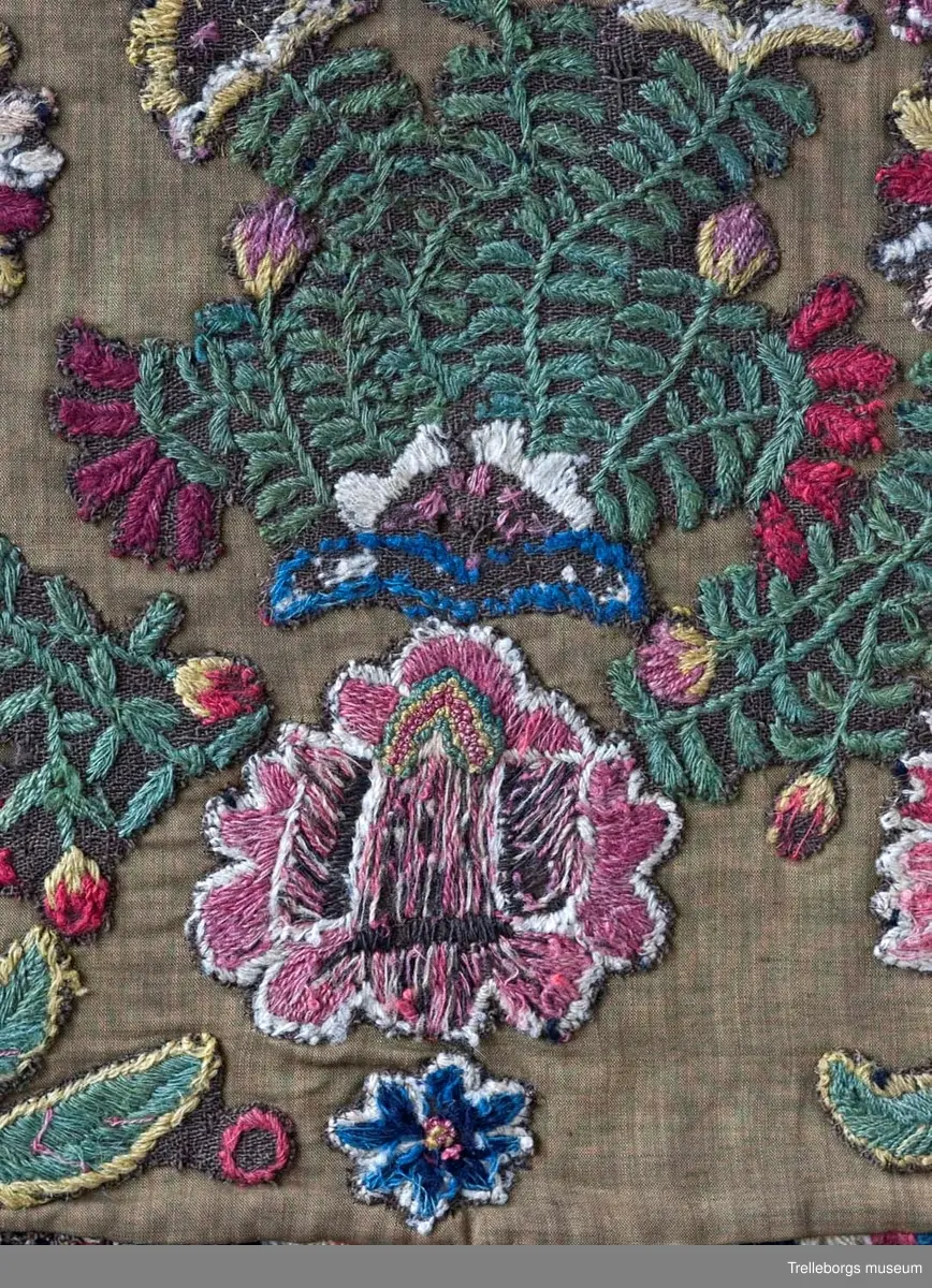 Broderad agedyna på grov, brun yllebotten. Motivet är två grönskande blomsterkransar fyllda med vad som mest liknar blommande buskar, även mellan kransarna finns två blommande buskar. Här är dynan märkt med CAD i den ena och ANO i den andra, båda på ett litet fält med franska knutar i vitt och rött. Under busken med ANO står 180 ganska glest, sista siffran finns inte kvar. Dynan är monterad med en flerfärgad kavelfrans och en klarröd, ganska grov yllebaksida.
