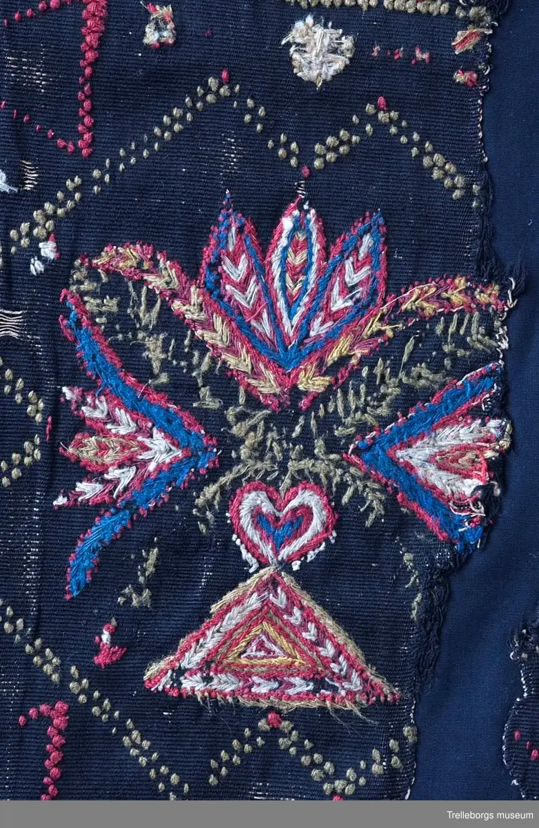 Material: varp 2 tr lingarn, inslag 1 tr ullgarn, ripsbindning. Broderi och flossaknutar 1 tr ullgarn. 
Bården runtom är vävd i tränsaflossa, diagonal inrutning. Dessutom är vävt konturer till tre rutor, dessa delar in agedynans mittfält i tre avdelningar. I tränsaflossan är även invävt konturerna till en hjärtfigur i vardera hörnet, samt två timglas utmed långsidorna. I var och en av de tre rutorna är broderat en blomstervas med tre tulpaner. Färgerna är här blått, cyklamen, gult, vitt och olivgrönt.

Tidigare konserverad då bitarna har sytts ihop på ny svart botten. Eventuellt kan det vara så att dynan klippts sönder för att brukas i två delar eller vid arvsskifte.